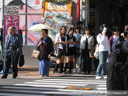 a parasol-toting woman in a kimono, some teenage girls, and others wait at a crosswalk in Kokura